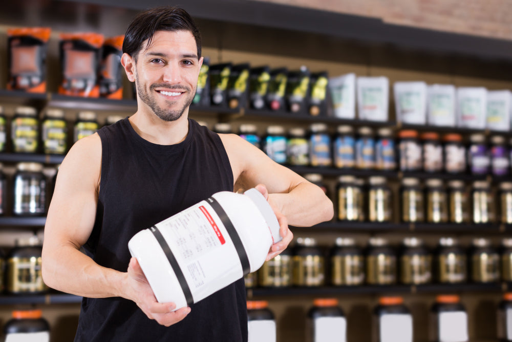 A smiling man in a sleeveless black shirt holds a large container of protein powder in a fitness supplement store, with various products displayed on shelves behind him.