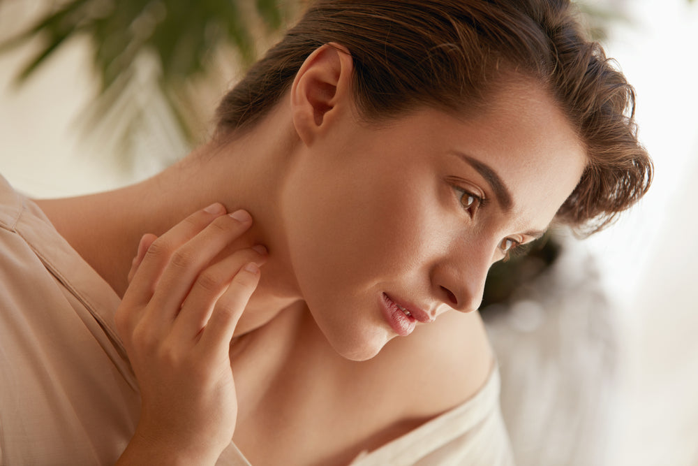 A woman with short, wavy hair gently touches her neck while gazing thoughtfully to the side, set against a soft, natural background that suggests a focus on beauty and wellness.