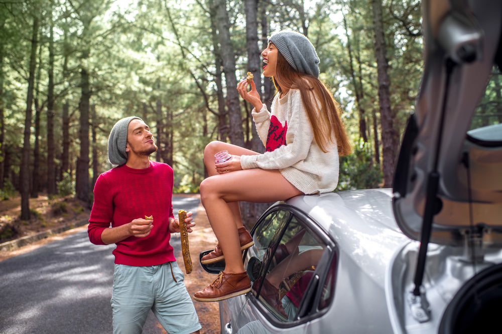 A man and woman enjoy keto snacks while sitting on a silver car parked on a forested road, with the woman laughing and the man smiling as they share the moment.