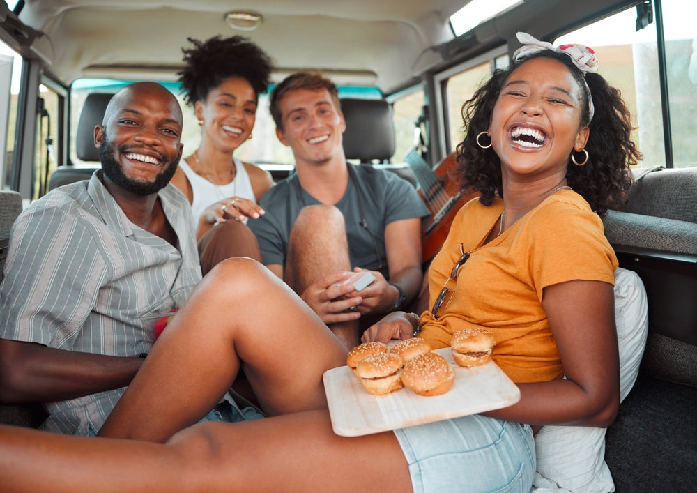 Four friends are sitting in a van, laughing and enjoying each other's company, while one holds a wooden platter with healthy snacks, creating a joyful road trip atmosphere.