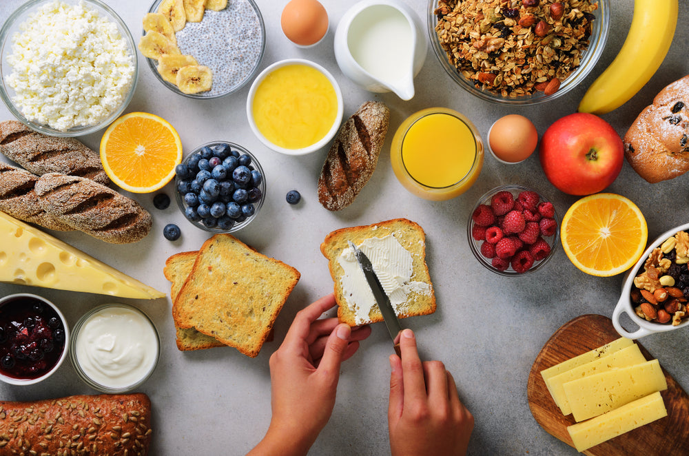 A hand spreads cream cheese on toasted bread surrounded by various breakfast items, including fruits, yogurt, granola, and drinks, arranged on a light gray surface.