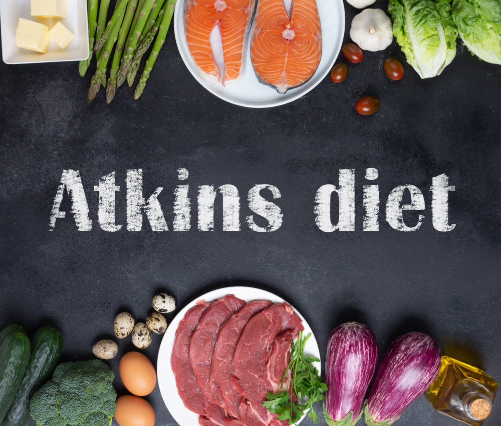 A variety of fresh foods including salmon, vegetables, eggs, and meat are arranged around the text "Atkins diet" on a dark surface, emphasizing low-carb dietary choices.
