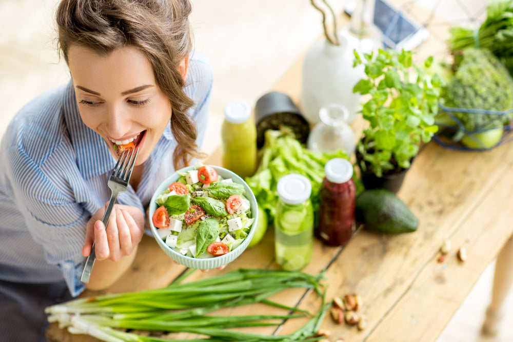 A woman in a striped shirt smiles as she takes a bite of salad from a bowl, surrounded by fresh vegetables and drinks on a wooden table in a bright, inviting kitchen.