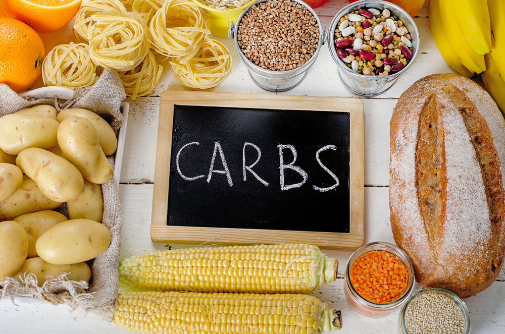 A variety of foods including potatoes, pasta, corn, fruits, and grains surround a chalkboard that reads "CARBS," highlighting different carbohydrate sources on a wooden table.