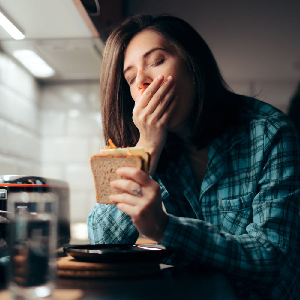A woman in a plaid shirt holds a sandwich while yawning, sitting at a kitchen counter with a glass of water and a plate nearby, suggesting she is tired during a meal.