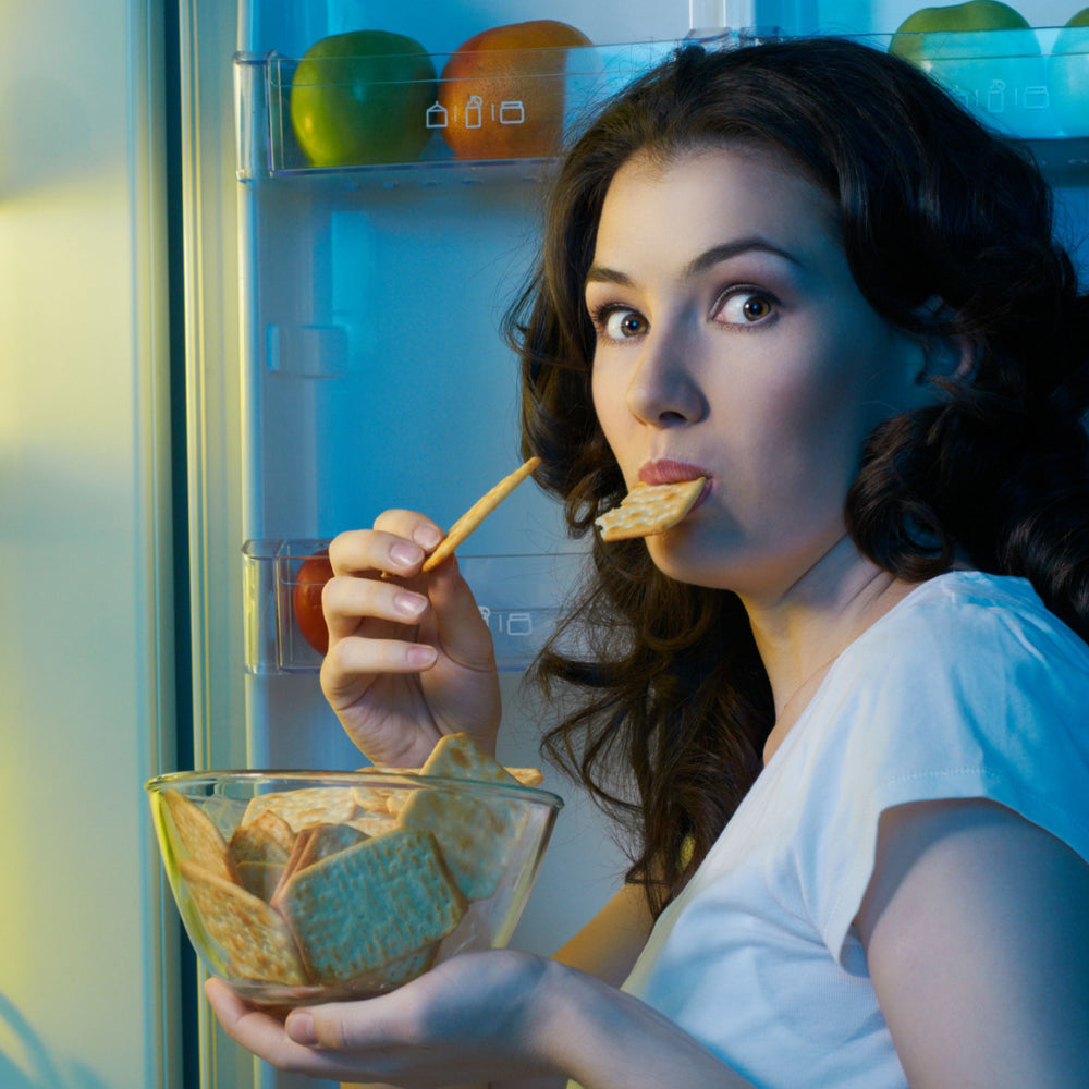 A woman with long, dark hair stands in front of an open refrigerator, holding a bowl of crackers in one hand and a cracker in her mouth, looking surprised or amused.
