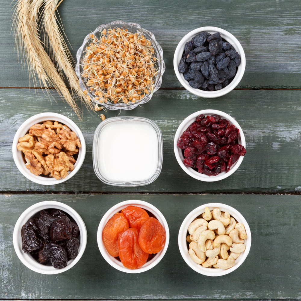 A variety of nuts, dried fruits, and sprouts are arranged in white bowls on a rustic wooden table, with a small container of milk in the center, showcasing healthy snack options.