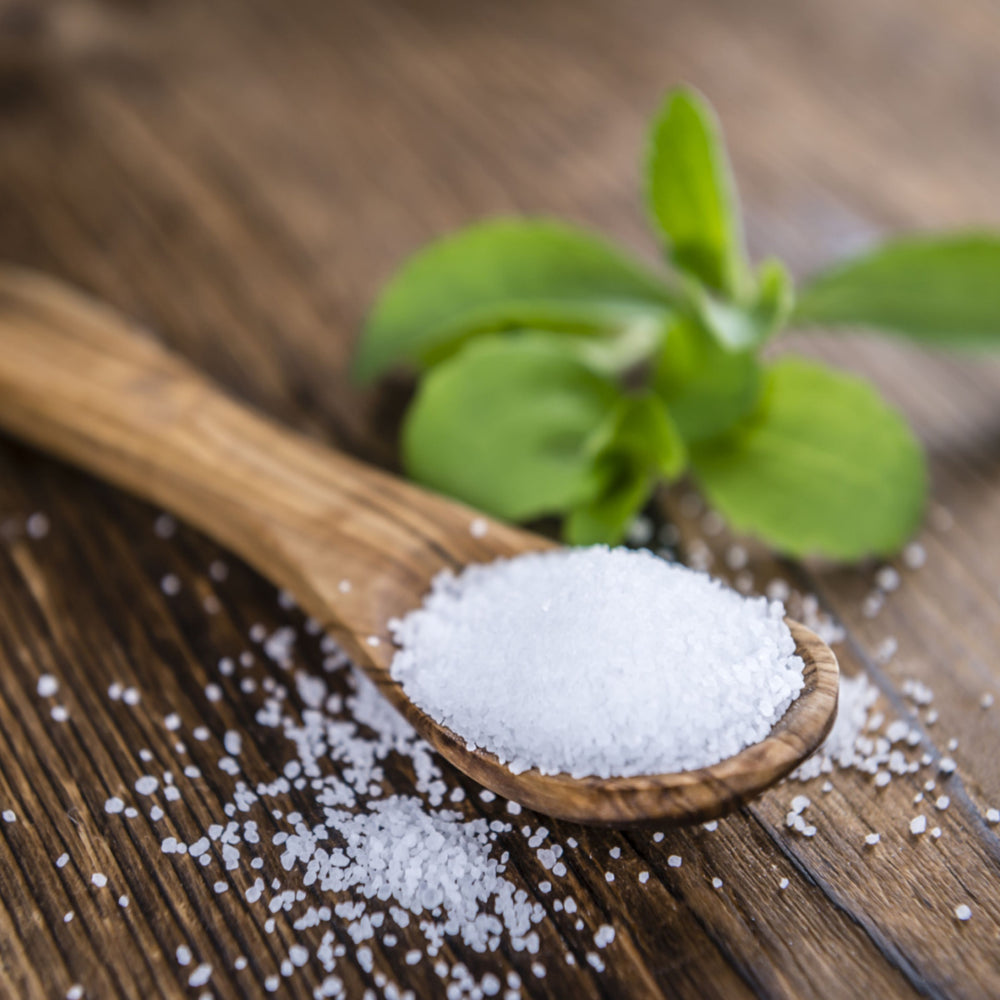 A wooden spoon holds white granulated salt, with some salt scattered on a wooden surface, alongside fresh green stevia leaves in the background.