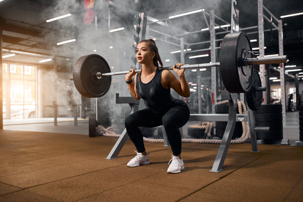 A woman in athletic wear squats with a barbell in a gym, focusing intently as she lifts the weight, surrounded by exercise equipment and a misty atmosphere.