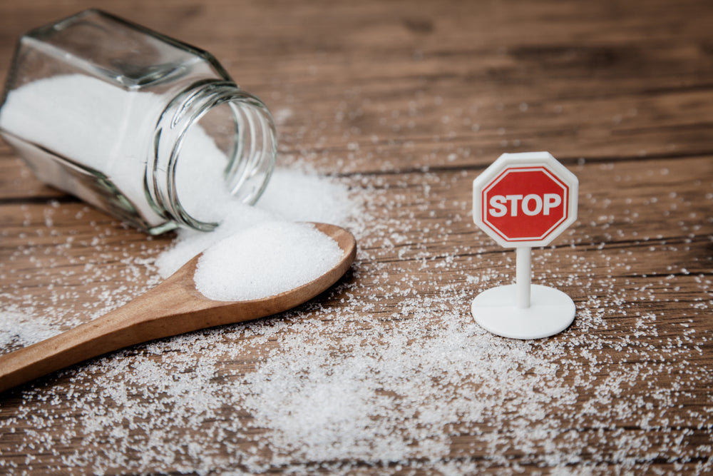 A glass jar of sugar spills onto a wooden surface, with a wooden spoon holding a scoop of sugar nearby and a small red stop sign placed in the spilled sugar, suggesting a cautionary message about sugar use.