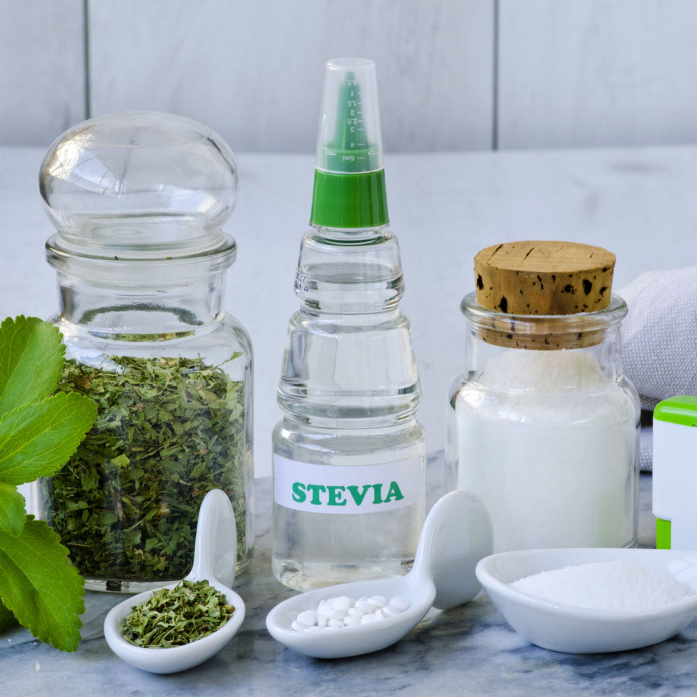 Various containers hold stevia products, including dried leaves, liquid extract, and sugar substitutes, arranged on a marble surface with a green plant leaf nearby.