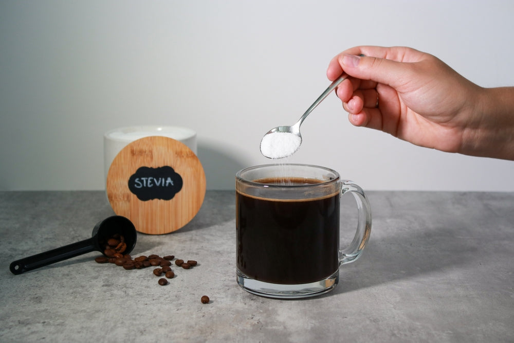A hand holds a spoonful of sweetener above a clear mug of black coffee, with a container labeled "Stevia" and scattered coffee beans in the background on a gray countertop.