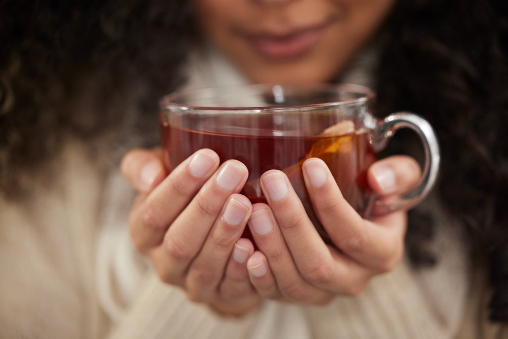 A person with curly hair holds a clear glass cup of tea close to their face, appearing to enjoy the warmth, while wearing a cozy sweater in a softly lit setting.