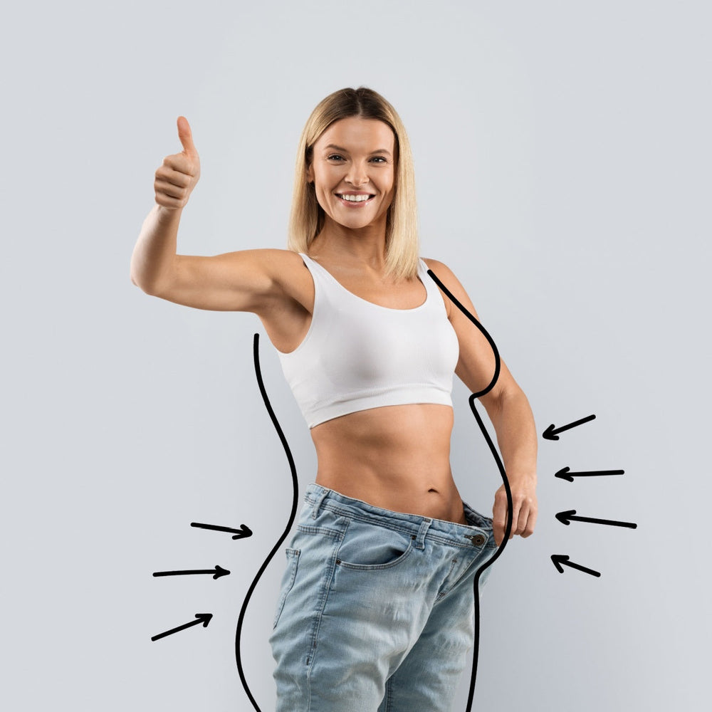 A smiling woman in a white tank top gives a thumbs-up while holding her oversized jeans, symbolizing successful weight loss and body transformation against a light gray background.