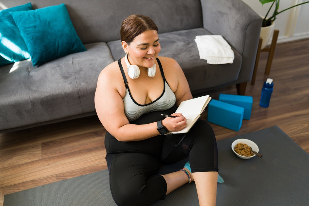 A smiling woman with headphones sits on a yoga mat, writing in a notebook, surrounded by a bowl of snacks, water bottle, and yoga blocks in a cozy living room setting.