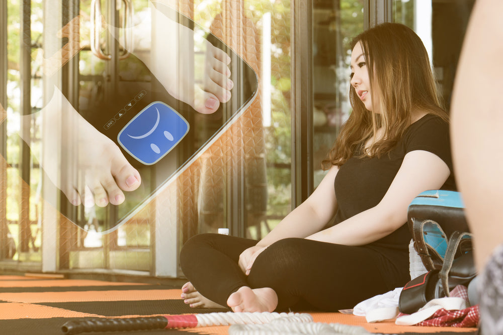 A woman sits on a gym mat, looking thoughtfully at a digital scale displaying her feet, with a bright, open fitness studio visible in the background.