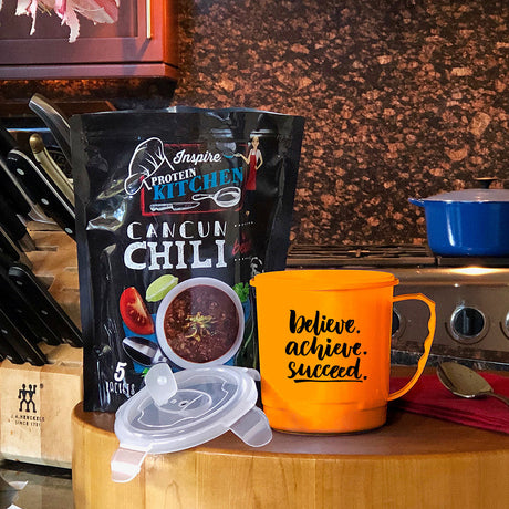 A black bag of "Inspire Protein Kitchen Cancun Chili" stands next to an orange mug with motivational text, on a wooden countertop in a cozy kitchen setting with cooking utensils in the background.