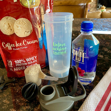 A clear shaker cup with motivational text sits on a countertop next to a bag of coffee-flavored protein powder, a scoop of powder, a bottle of Fiji water, and a shaker lid.
