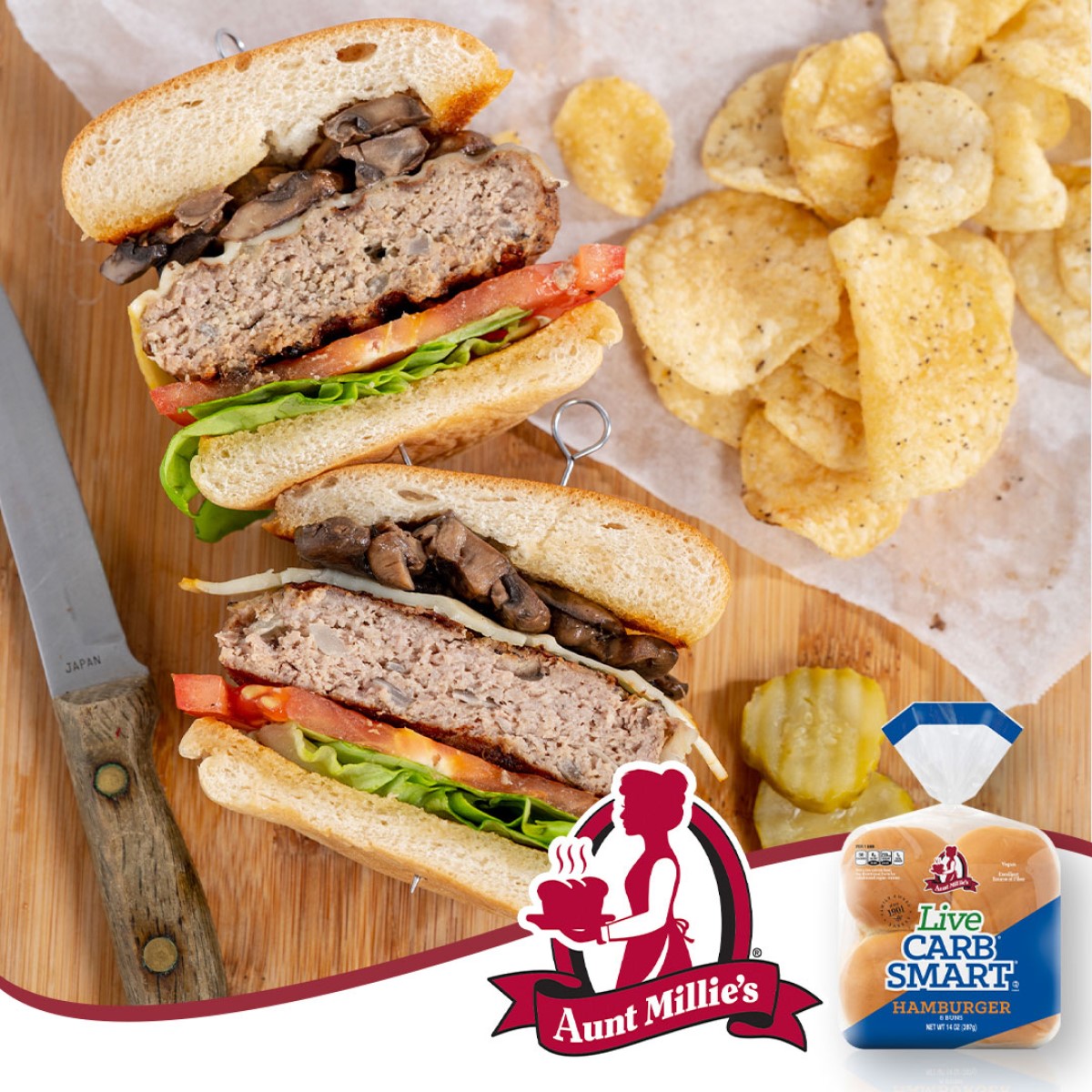 A close-up of a sliced hamburger stacked with lettuce, tomato, and mushrooms, served with potato chips on a wooden board, alongside Aunt Millie's Carb-Smart hamburger buns and a knife.