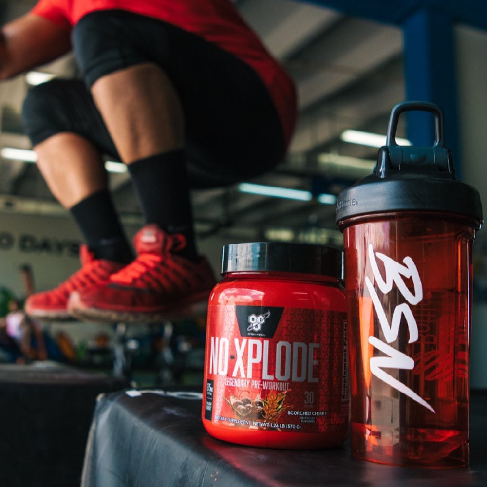 A person in a red shirt performs a squat in a gym, with a jar of BSN NO X-PLODE pre-workout and a shaker bottle visible on a black surface nearby.