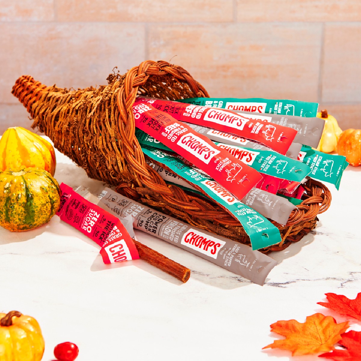 A cornucopia filled with colorful Chomps meat sticks sits on a marble surface, surrounded by small pumpkins and autumn leaves, creating a festive fall atmosphere.