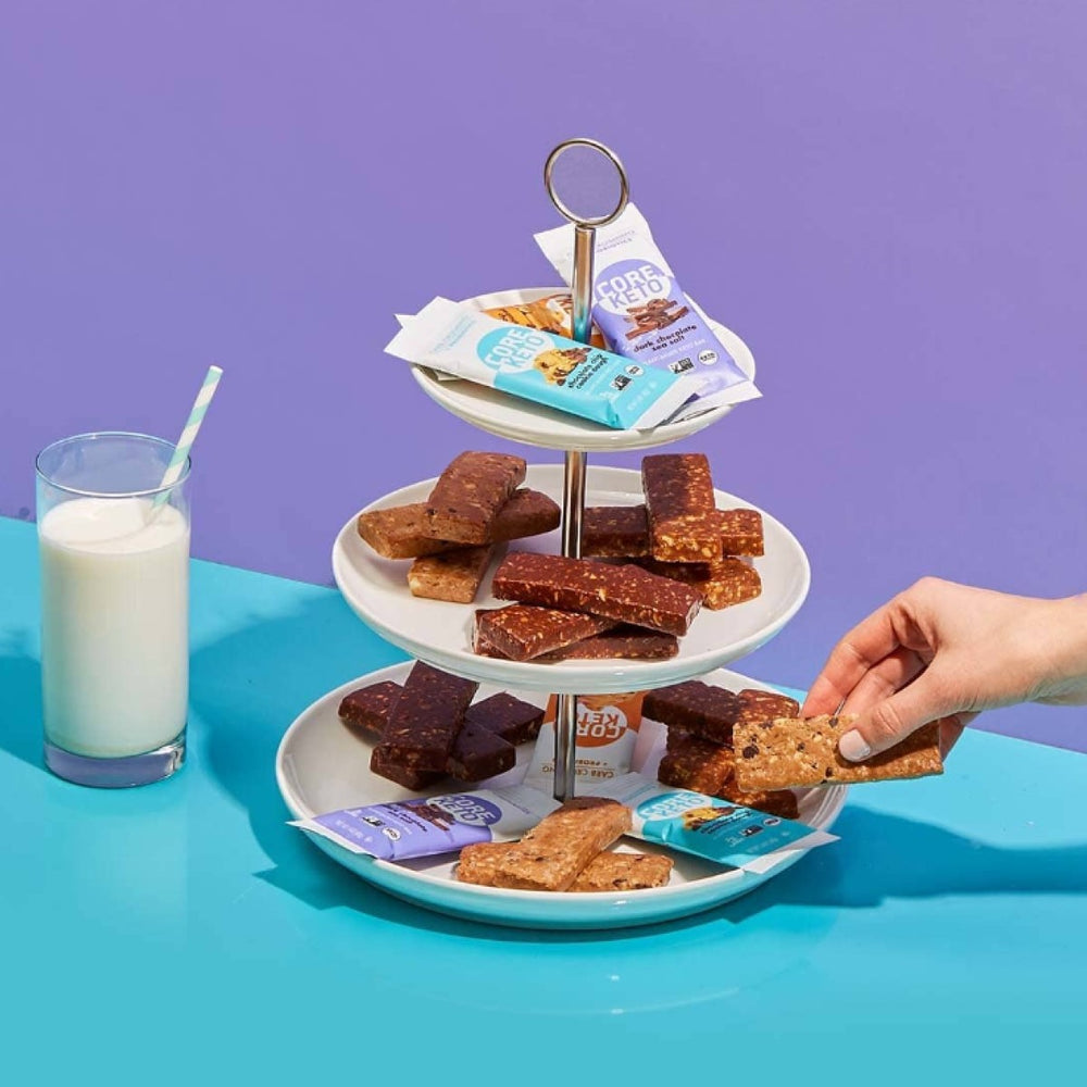 A hand reaches for a snack bar from a three-tiered stand filled with various snack bars, next to a glass of milk with a straw, set against a purple background.