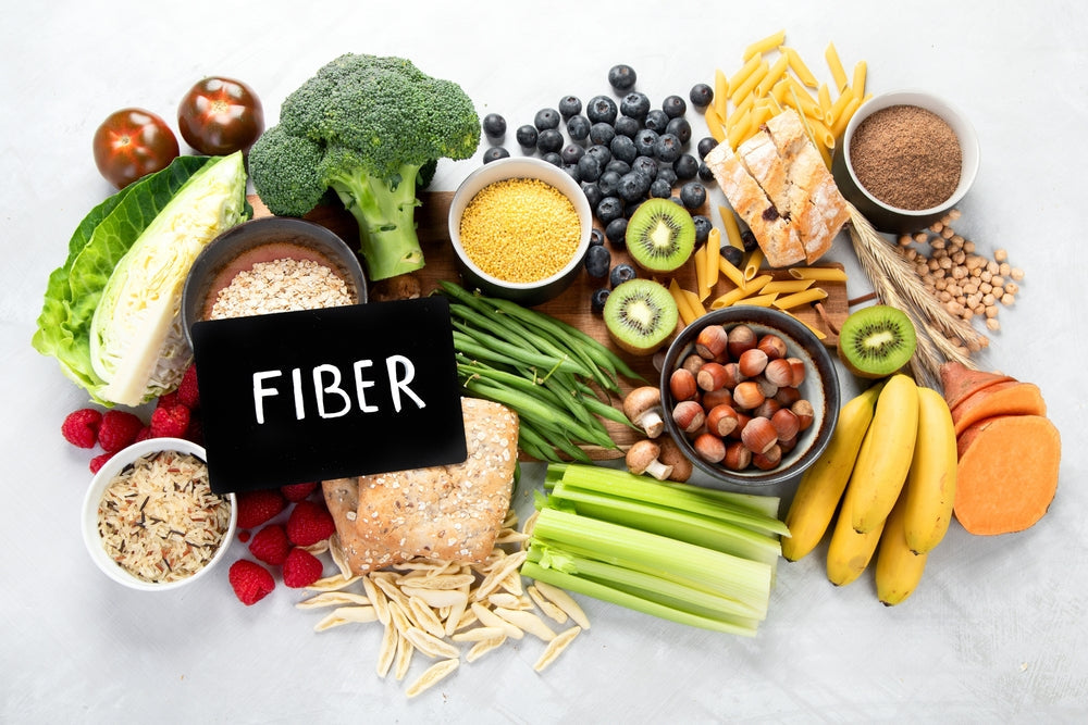 A variety of colorful foods rich in fiber, including broccoli, bananas, oats, and beans, are arranged on a light surface, with a black sign labeled "FIBER" prominently displayed.