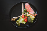 A black plate holds a piece of raw salmon, cherry tomatoes, broccoli, lettuce, and a quail egg, accompanied by wooden chopsticks and a lemon wedge, set against a dark wooden background.