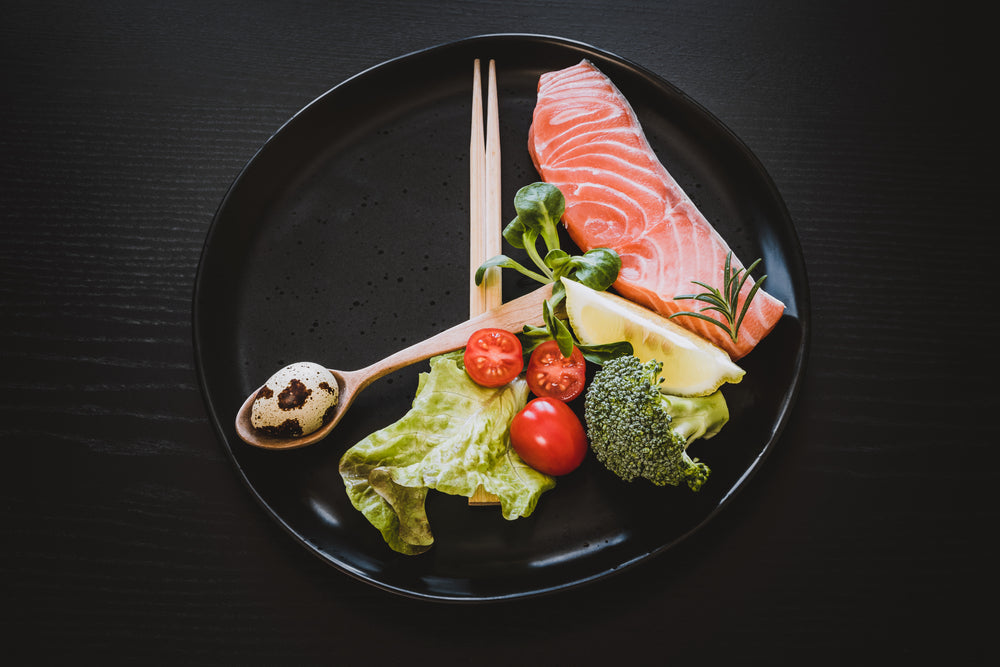 A black plate holds a piece of raw salmon, cherry tomatoes, broccoli, lettuce, and a quail egg, accompanied by wooden chopsticks and a lemon wedge, set against a dark wooden background.
