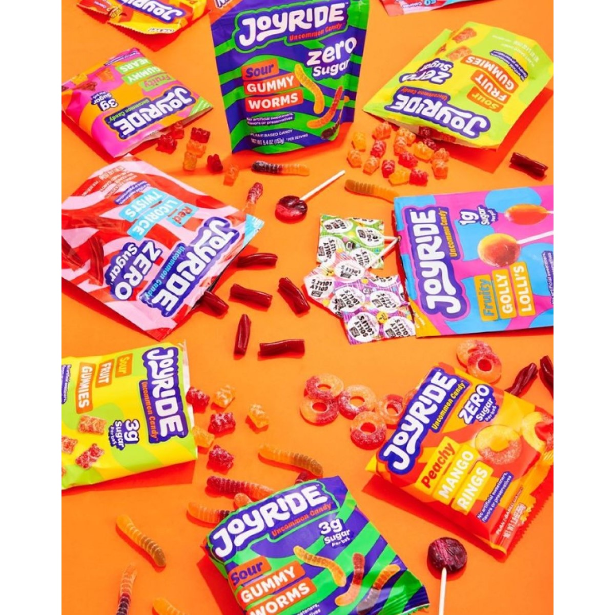 Colorful packages of Joyride candy, including gummy worms and lollipops, are scattered on a bright orange background, surrounded by various candies and wrappers.