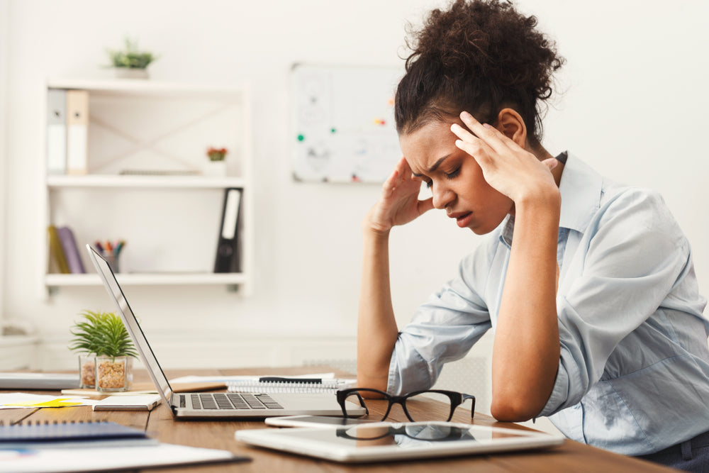 A woman sits at a desk with a laptop, looking stressed and holding her head, in a bright office setting with plants and notebooks around her, suggesting she is experiencing symptoms related to keto flu.