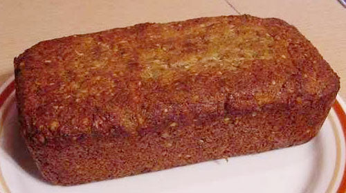 A freshly baked loaf of low carb zucchini coconut pecan bread sits on a round plate, showcasing a golden-brown crust and a moist texture, ready to be sliced and served.