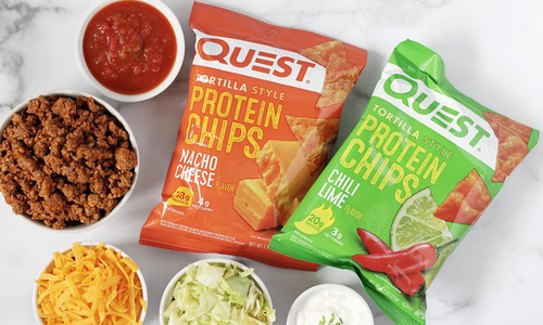 Two bags of Quest protein chips in nacho cheese and chili lime flavors are surrounded by bowls of seasoned ground meat, shredded cheese, lettuce, sour cream, and salsa on a white surface.