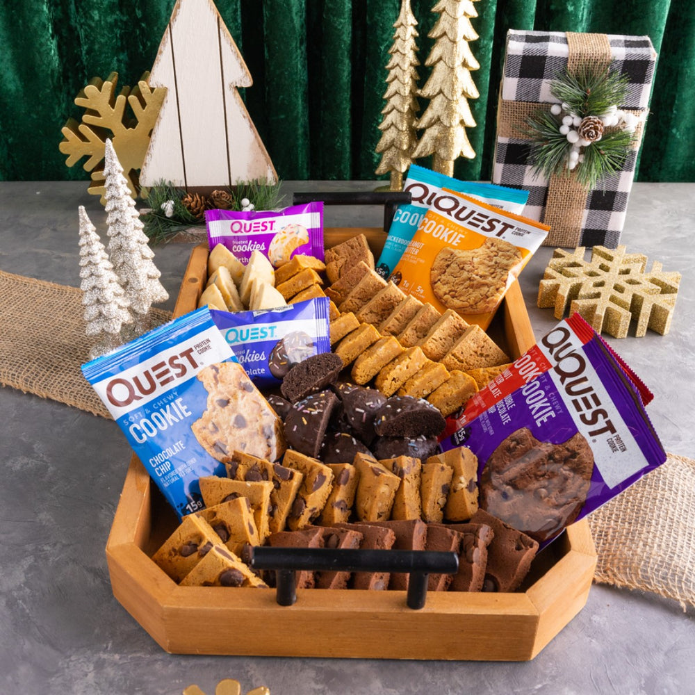 A wooden tray filled with various Quest cookies and snack bars is surrounded by festive decorations, including small trees, a wrapped gift, and a burlap runner on a gray surface.