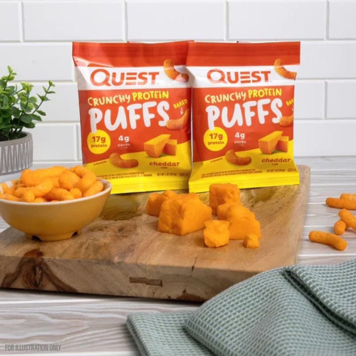Two bags of Quest Crunchy Protein Puffs are displayed on a wooden cutting board alongside a bowl of cheese puffs and cheddar cheese cubes, with a green kitchen towel in the foreground.