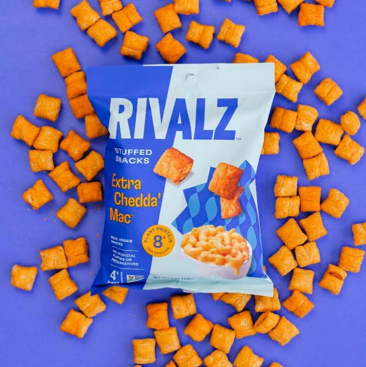 A bag of Rivalz Extra Chedda' Mac stuffed snacks is displayed on a vibrant purple background, surrounded by scattered orange snack pieces, highlighting the product's playful and colorful branding.