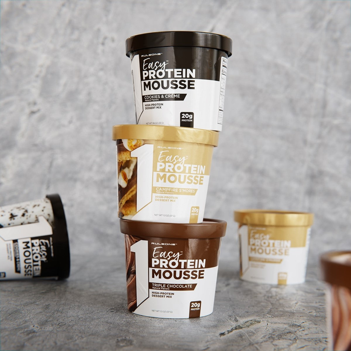 Three tubs of Easy Protein Mousse are stacked, featuring flavors Cookies & Crème, Campfire S'mores, and Triple Chocolate, against a textured gray background.