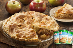 A freshly baked apple pie sits in a glass dish with a slice removed, revealing its warm, spiced filling, surrounded by whole apples and cans of Comstock apple filling on a rustic wooden table.