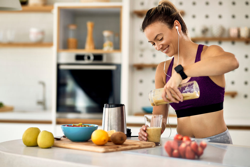 A woman in a purple sports top pours a smoothie from a blender into a glass while smiling, surrounded by fresh fruits and a modern kitchen setting.