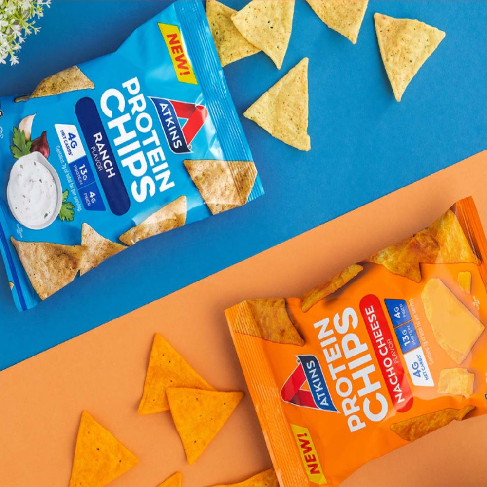 Two bags of Atkins Protein Chips, one ranch-flavored and one nacho cheese-flavored, are displayed on a blue and orange background, surrounded by scattered triangular chips.