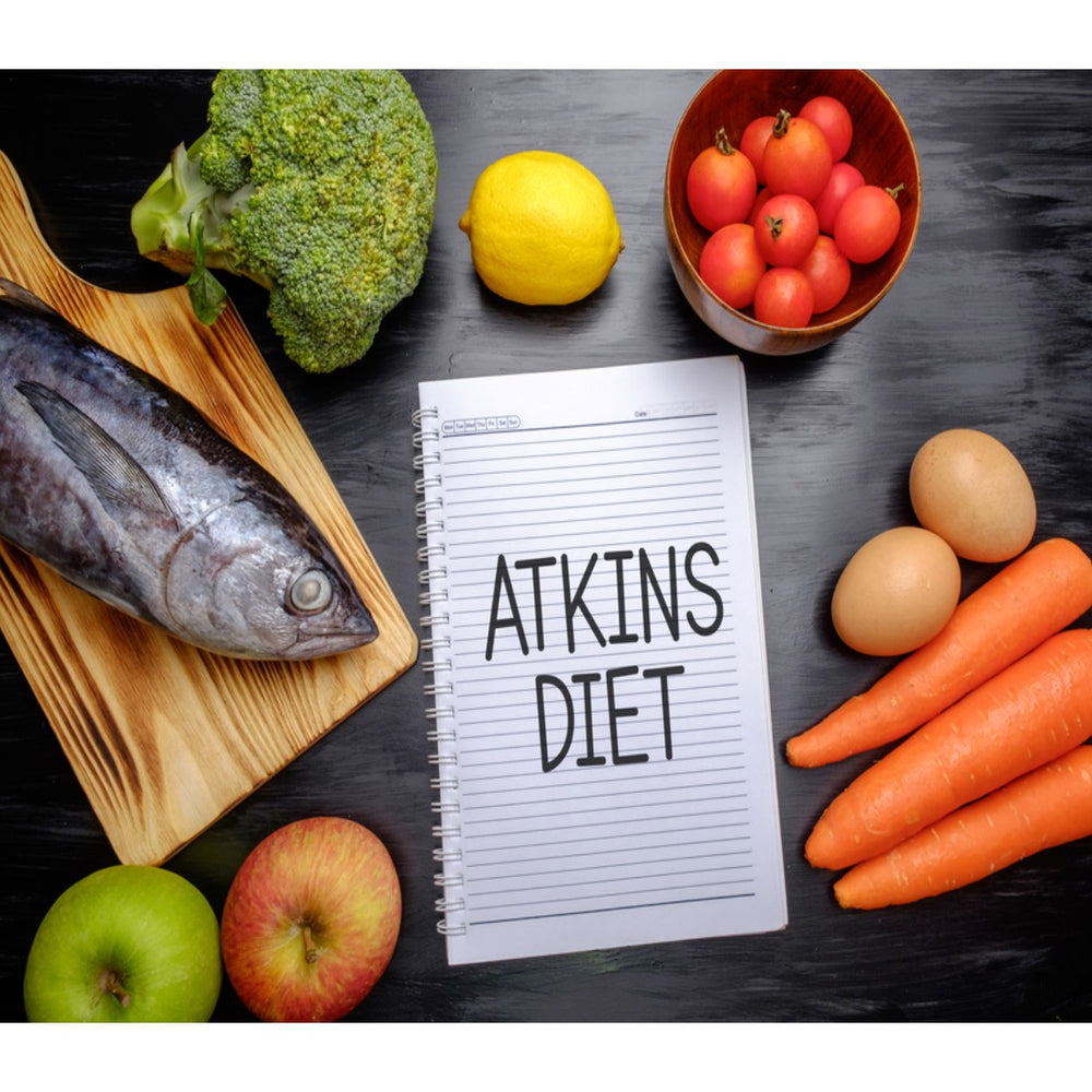 A variety of fresh foods, including a whole fish, broccoli, lemon, tomatoes, apples, carrots, and eggs, surround a notebook labeled "Atkins Diet" on a dark surface.