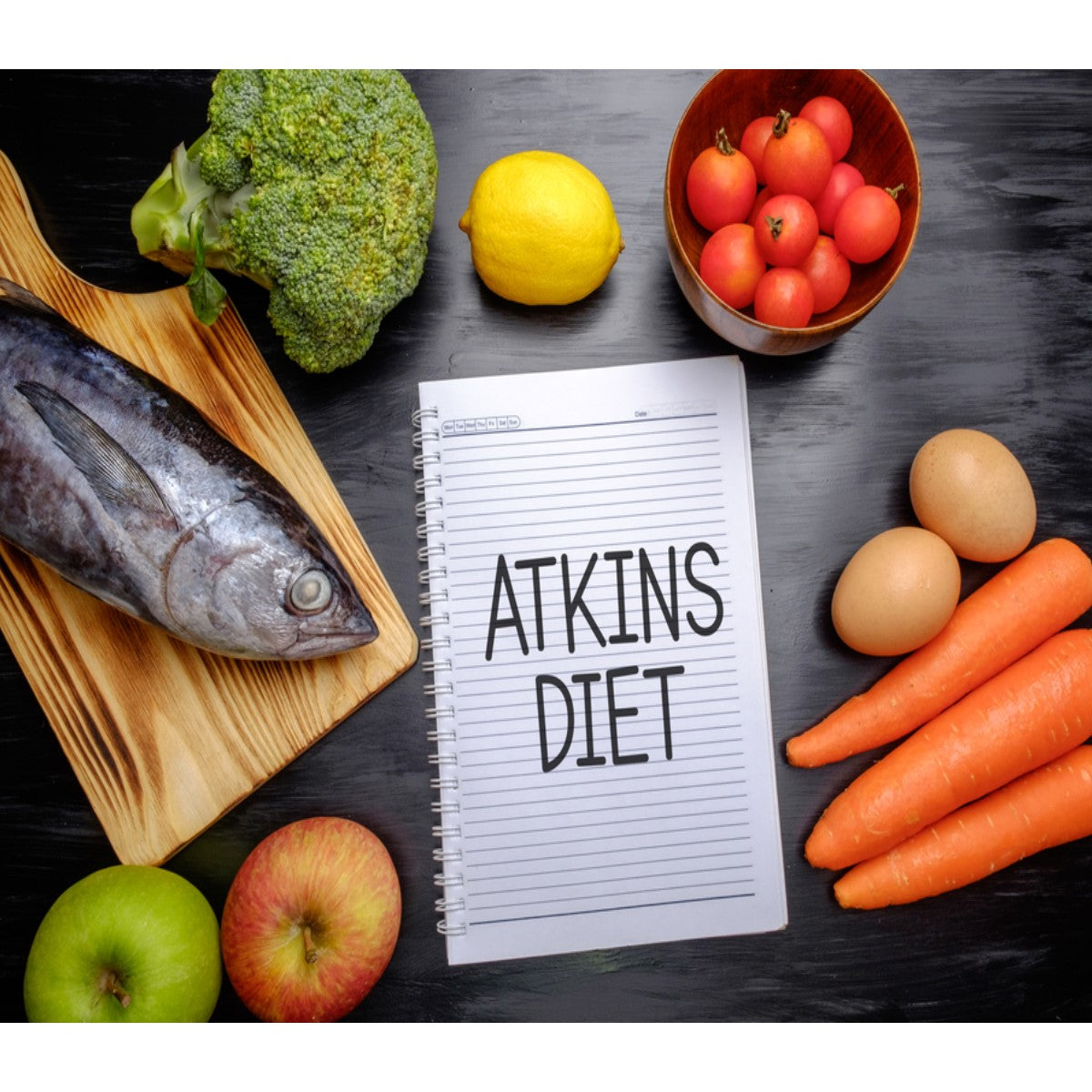 A variety of fresh foods, including a whole fish, broccoli, lemon, tomatoes, apples, carrots, and eggs, surround a notebook labeled "Atkins Diet" on a dark surface.