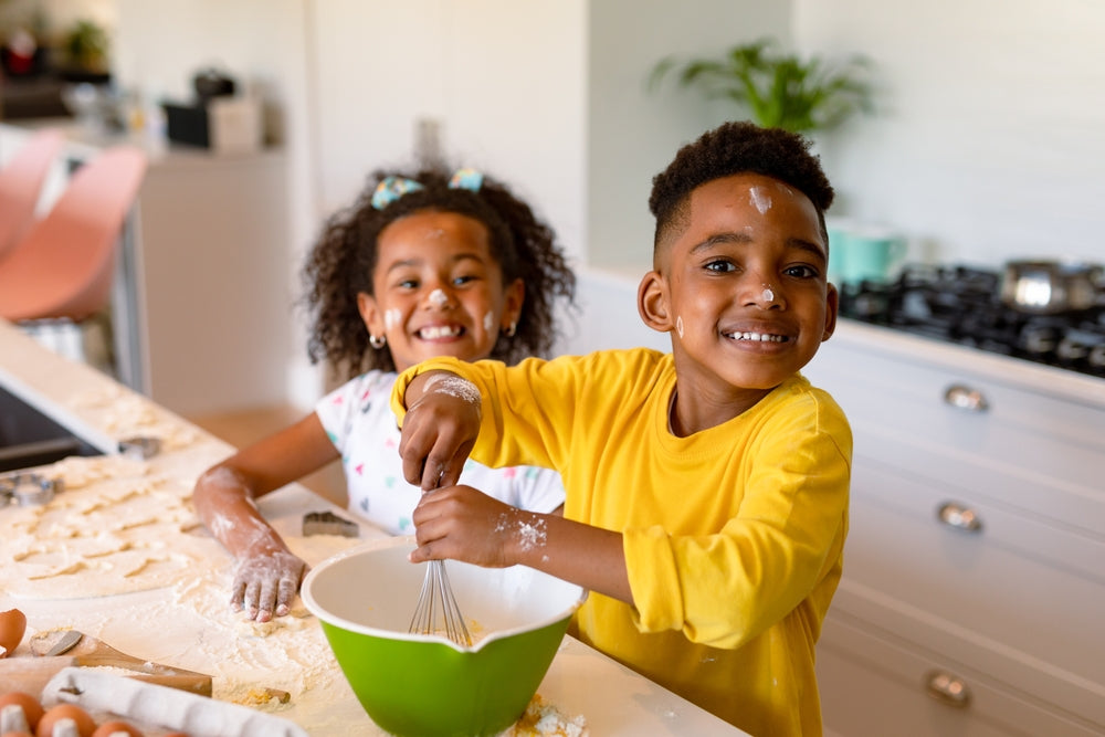 Two smiling children, a girl with curly hair and a boy in a yellow shirt, playfully mix ingredients in a green bowl while surrounded by flour and baking supplies in a bright kitchen.