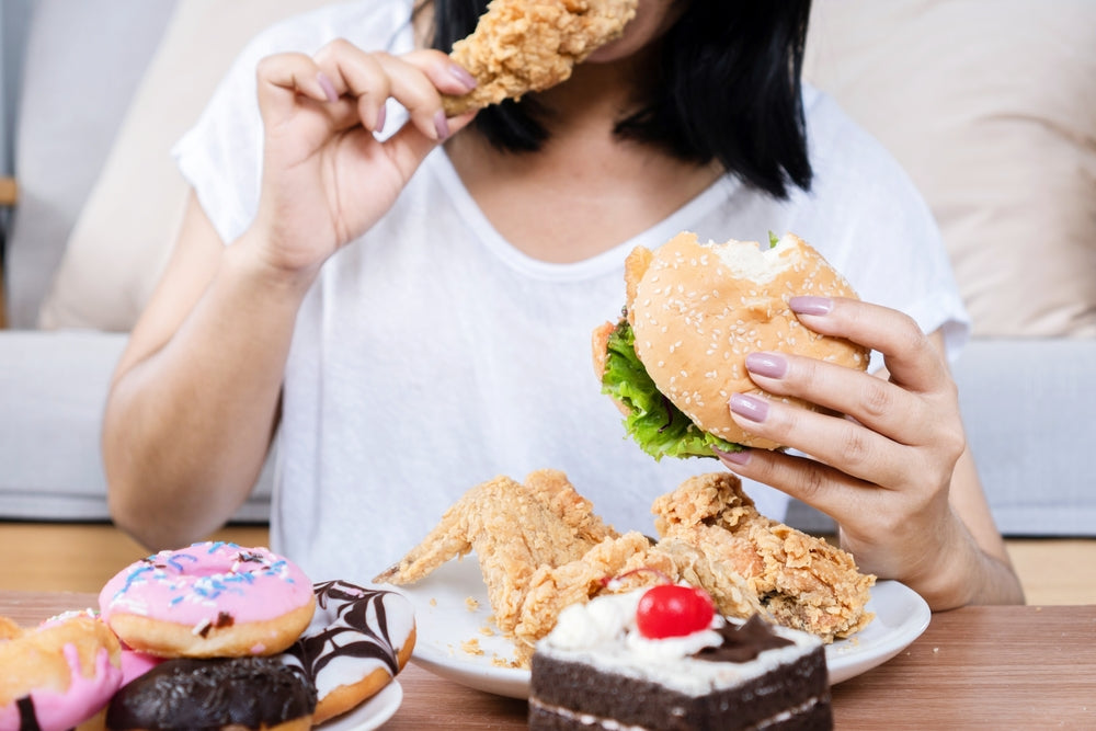 A person in a white shirt sits on the floor, holding a piece of fried chicken in one hand and a burger in the other, surrounded by donuts and cake on a plate, suggesting a moment of indulgence.