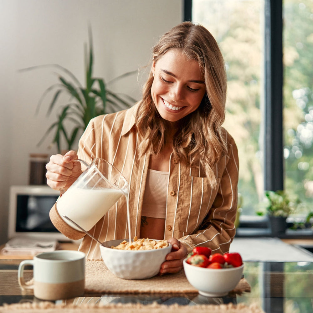 A smiling woman in a striped shirt pours milk into a bowl of cereal at a table, with a cup and a bowl of strawberries nearby, surrounded by greenery and natural light.