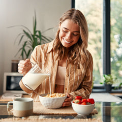 A smiling woman in a striped shirt pours milk into a bowl of cereal at a table, with a cup and a bowl of strawberries nearby, surrounded by greenery and natural light.