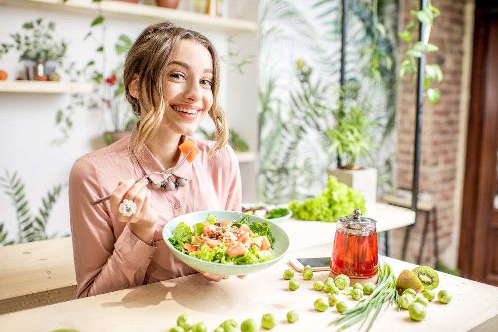 A smiling woman in a pink blouse holds a bowl of colorful salad while sitting at a wooden table adorned with fresh vegetables and a glass of tea, surrounded by greenery in a bright, modern setting.