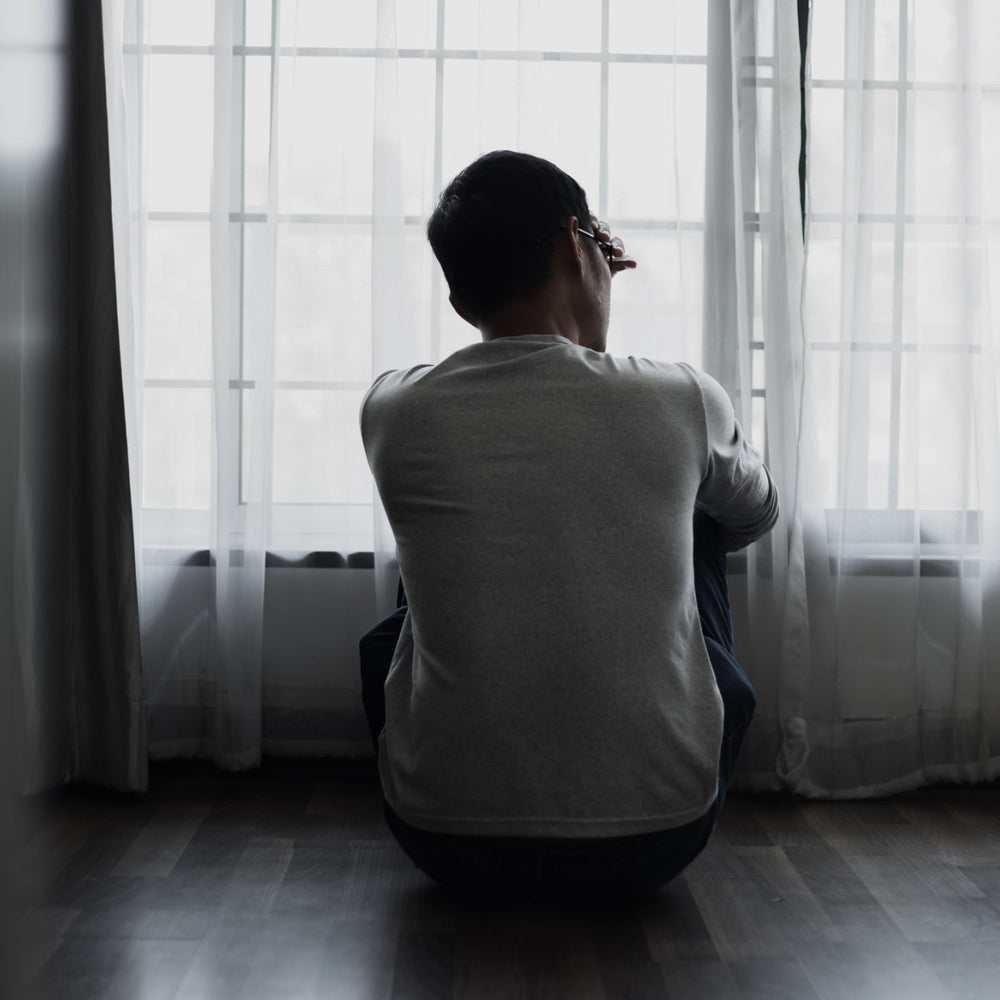 A person sits on the floor, facing away from the camera, with their knees drawn up and head resting on their hand, gazing out through sheer curtains at a bright, overcast window.