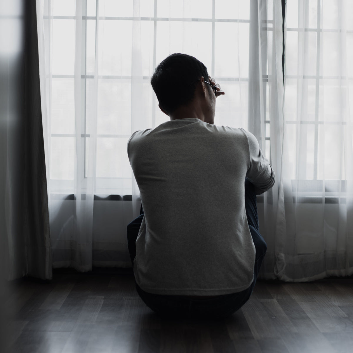 A person sits on the floor, facing away from the camera, with their knees drawn up and head resting on their hand, gazing out through sheer curtains at a bright, overcast window.