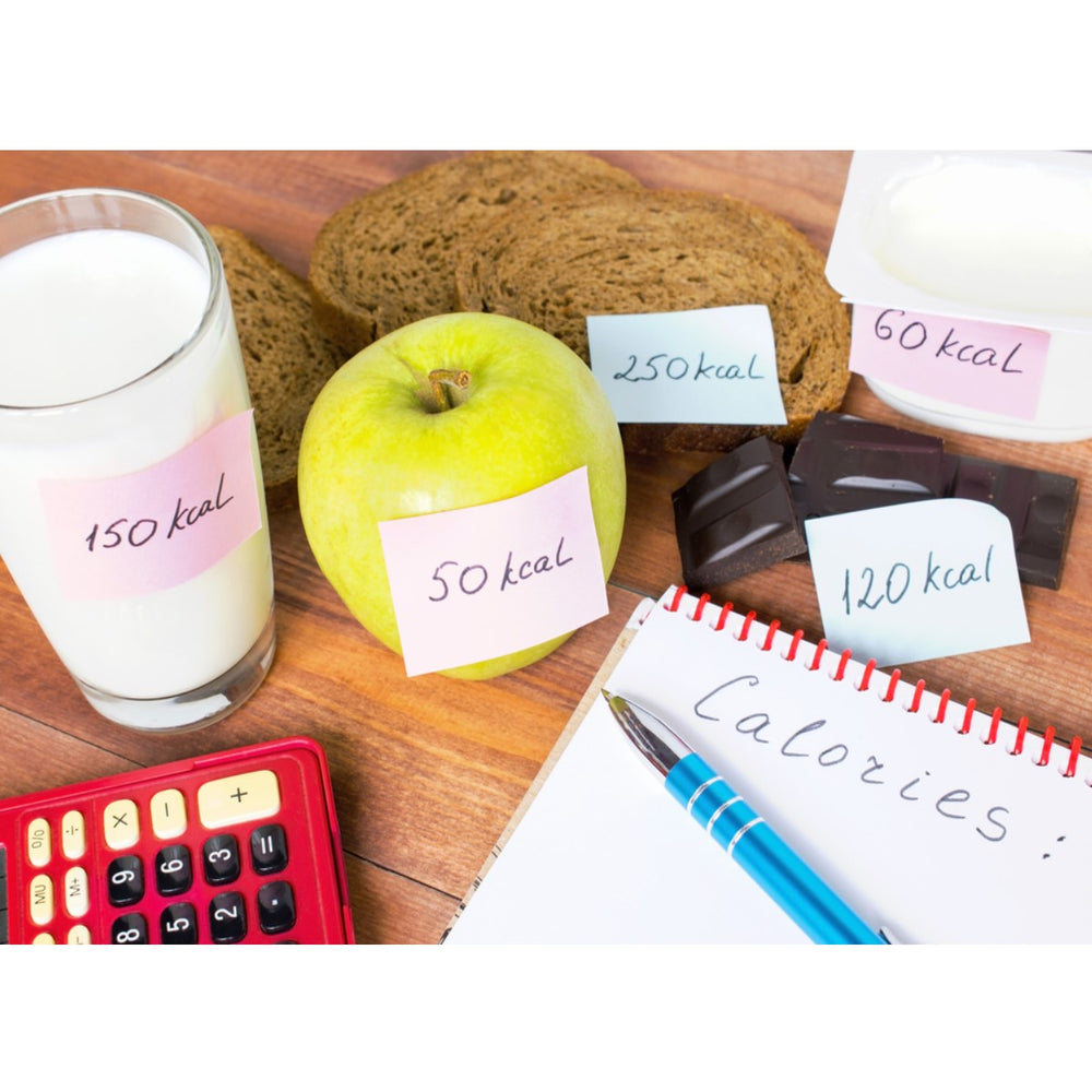 A glass of milk, a green apple, slices of brown bread, chocolate pieces, and yogurt are labeled with calorie counts, alongside a notepad and calculator on a wooden table.